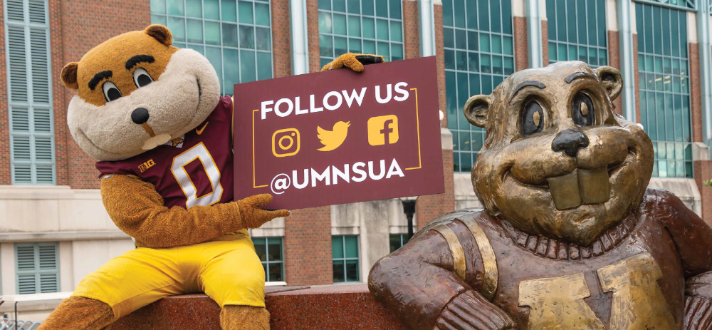 Goldy Gopher sitting near Goldy statue outside Coffman Memorial Union holding a sign that states: Follow Us #UMNSUA with social media icons for instagram, Twitter, and facebook