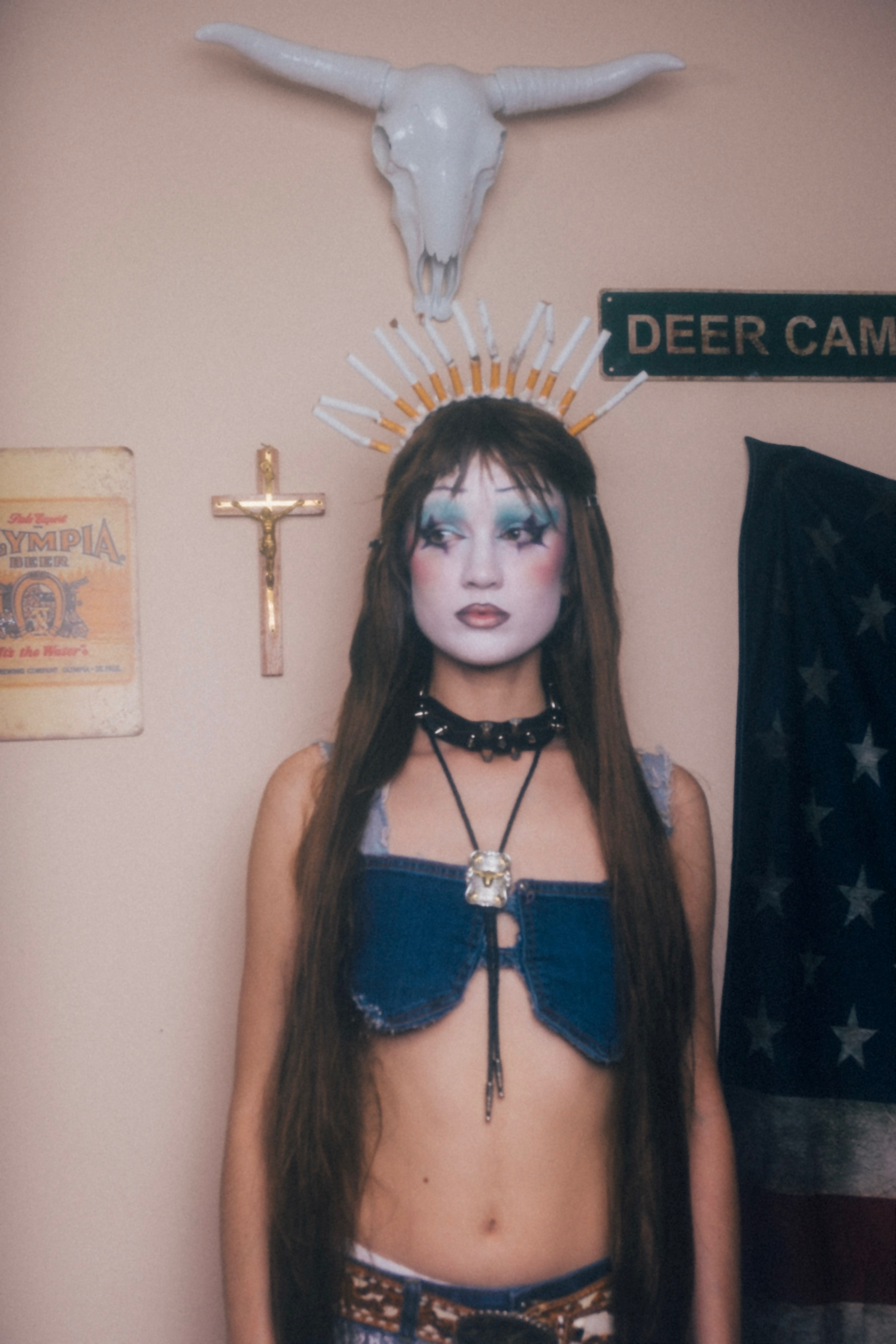 Singer Susannah Joffe standing underneath a longhorn skull and crucifix, wearing a crown of cigarettes on their head. 