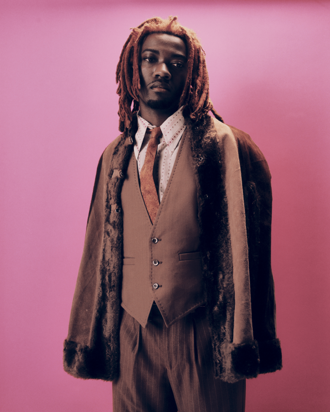 Portrait of a person in a brown tailored outfit and fur-trimmed coat standing against a pink background.